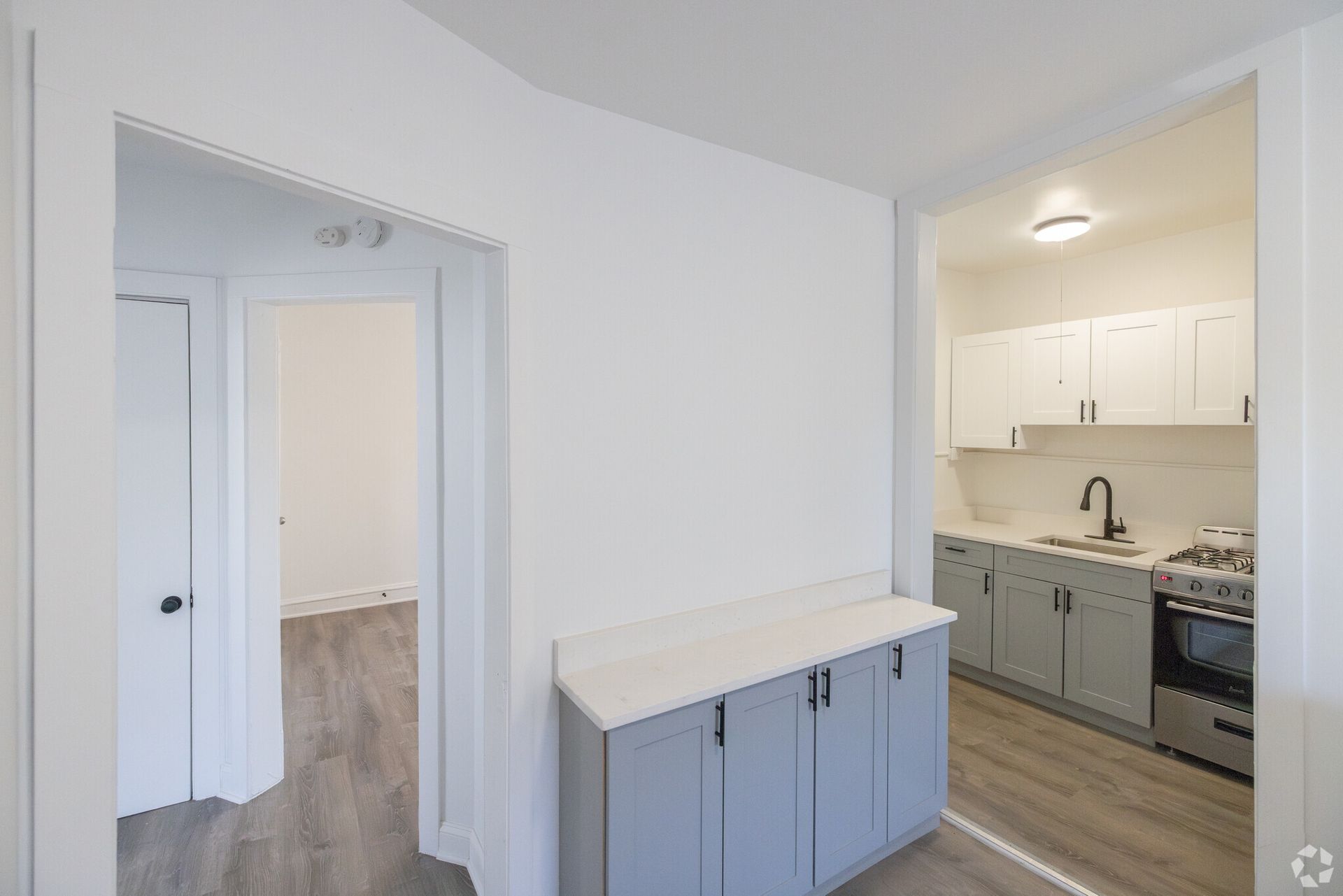 Interior view: hallway, door to bedroom, kitchen with gray cabinets, white walls, and countertop.