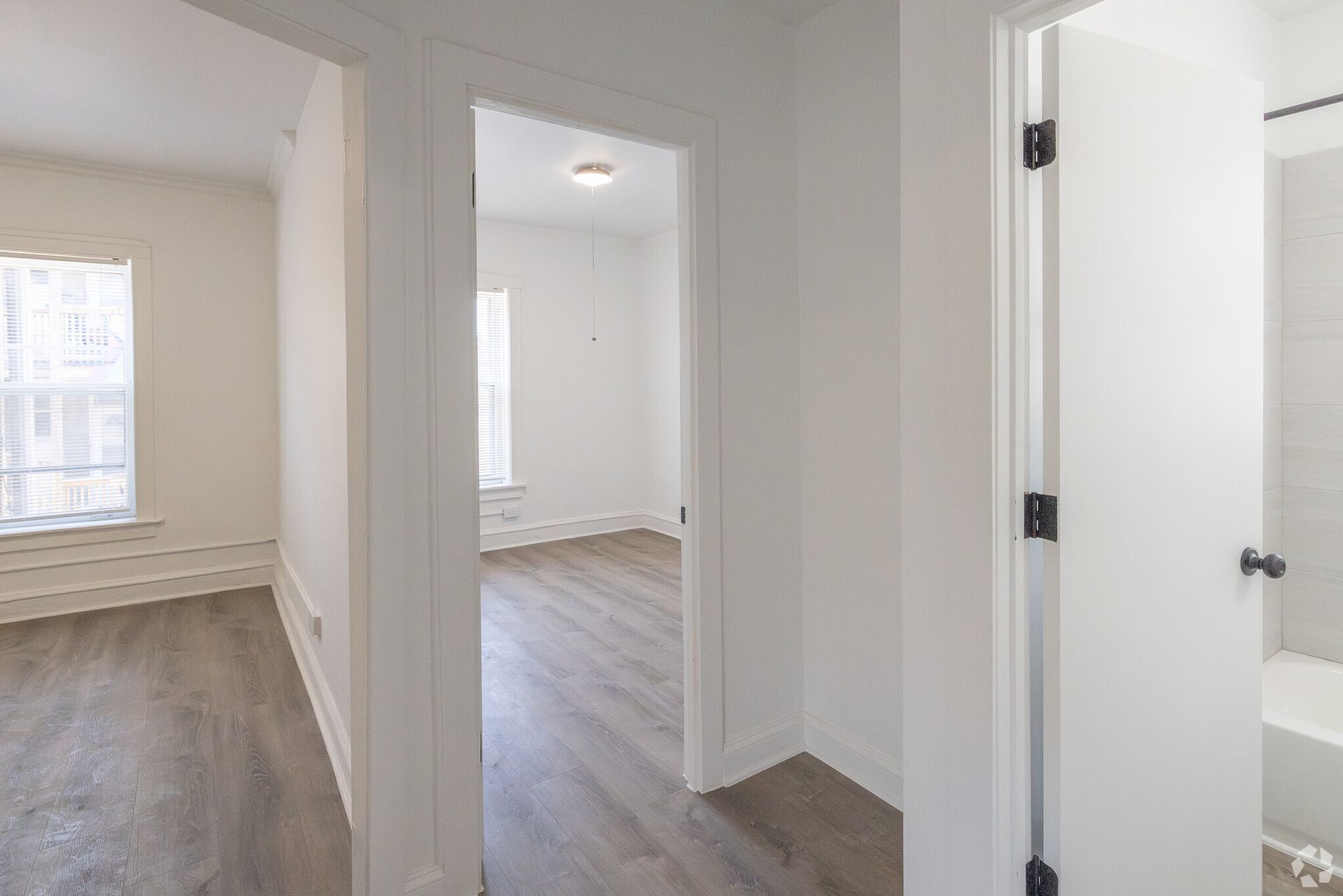 Hallway with open doorways to rooms and a bathroom, all with white walls and light wood floors.