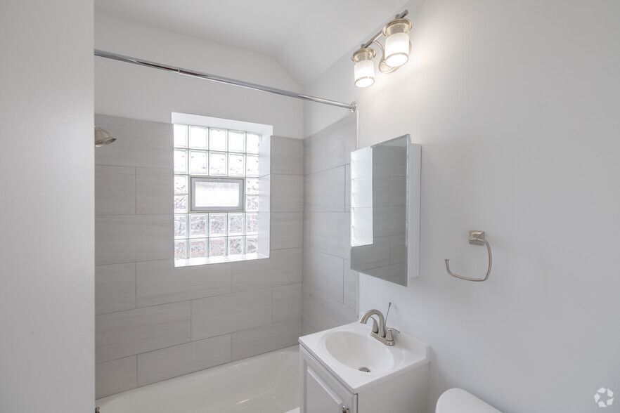 White bathroom with light gray tile in shower area, vanity, mirror, and glass block window.