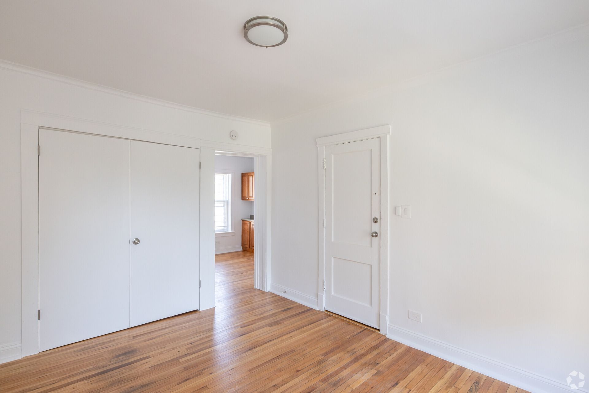 Empty room with hardwood floors, white walls, closet, and door leading to a kitchen.