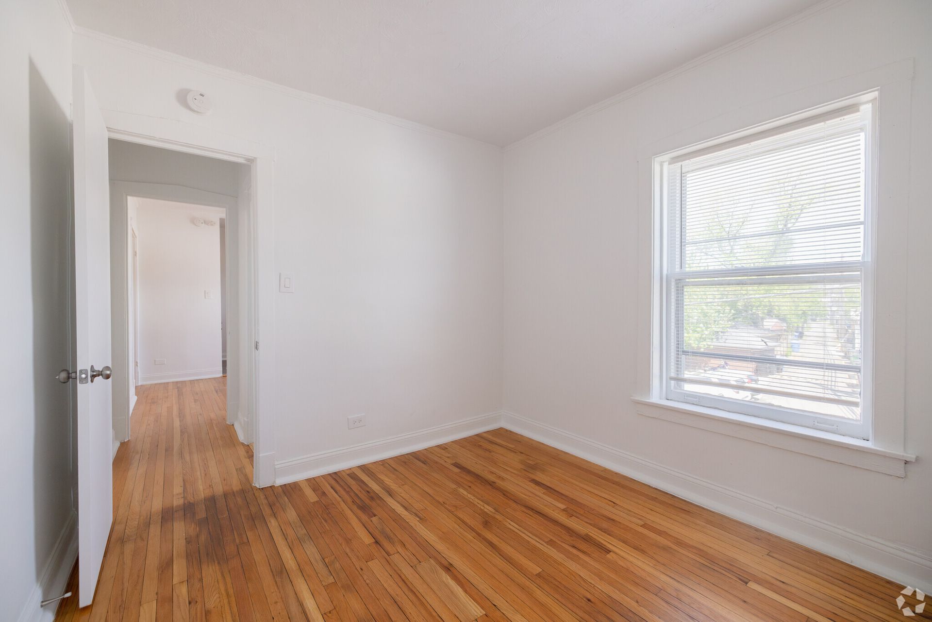 Empty room with hardwood floors, window, and doorway to a hallway. White walls.