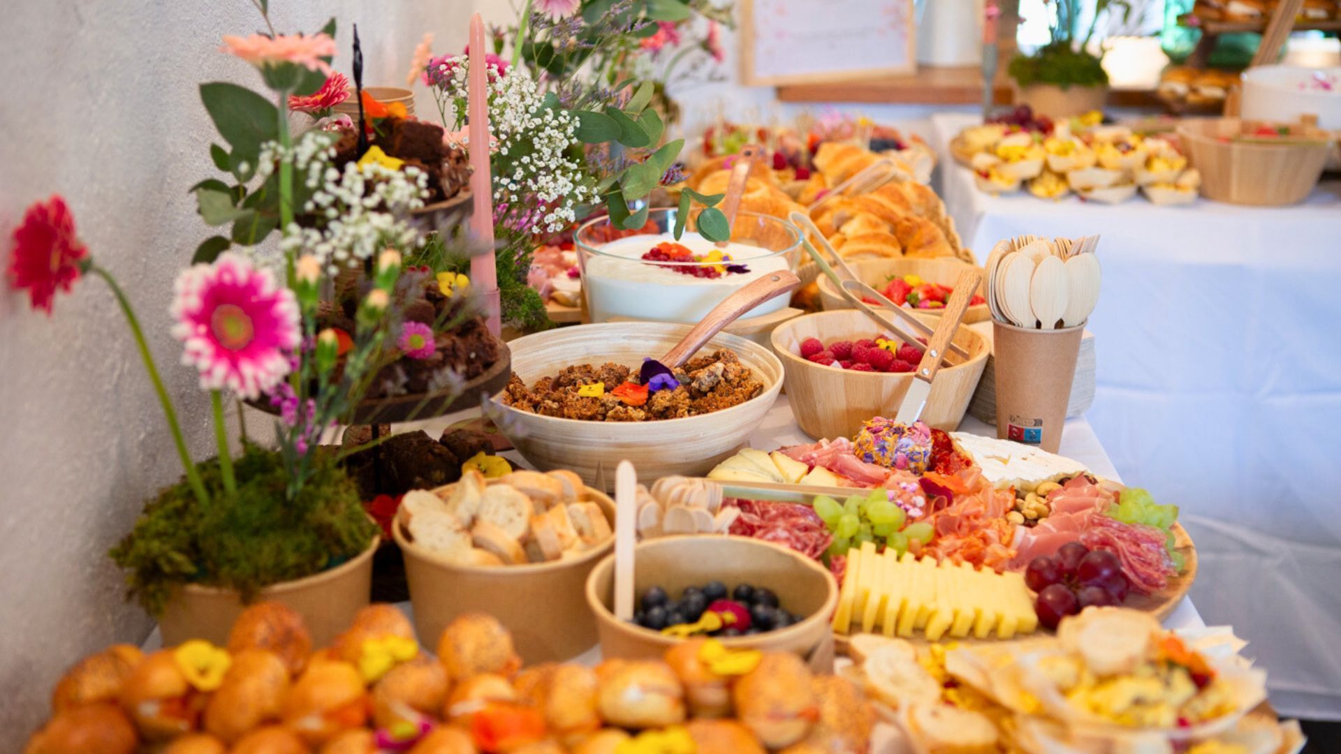 Un buffet de petit-déjeuner garni de bols de granola, de fruits, de viennoiseries et de charcuterie, décoré de fleurs fraîches.