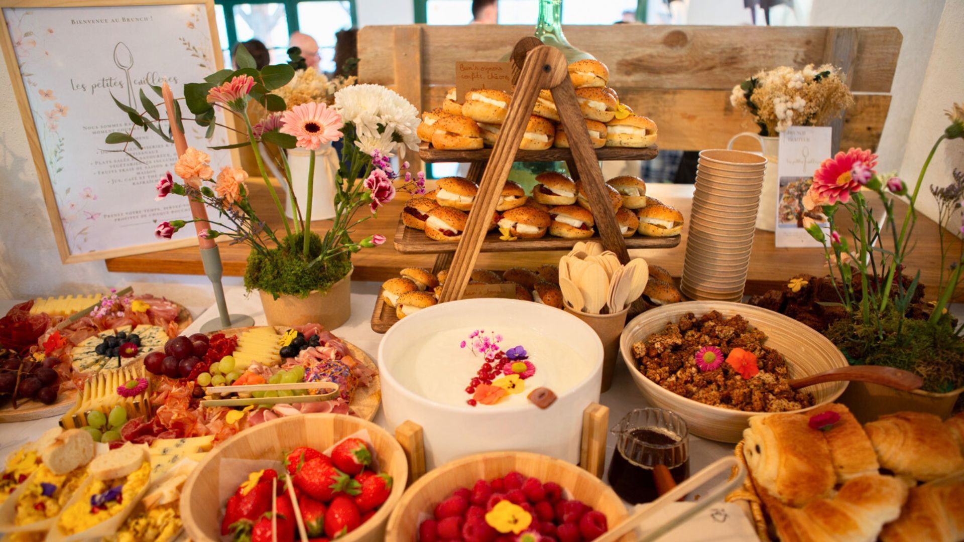 Une table de banquet garnie de pâtisseries assorties, de fruits frais, de granola, de charcuterie savoureuse et de centres de table floraux.