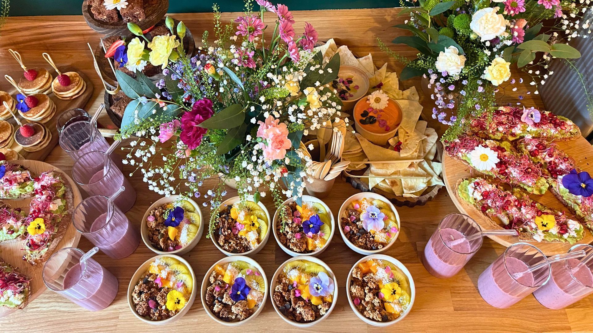 Une table en bois dressée avec des bols de nourriture recouverts de fleurs, des toasts à l'avocat et des petites boissons roses, décorée de bouquets de fleurs fraîches.