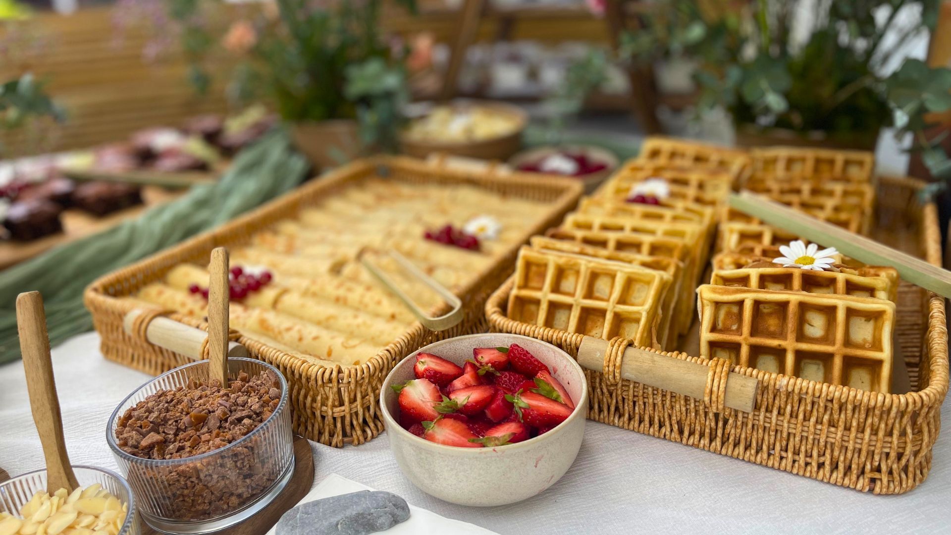 Un buffet de petit-déjeuner comprenant un panier de gaufres, un plateau de crêpes roulées, un bol de fraises coupées et des garnitures.