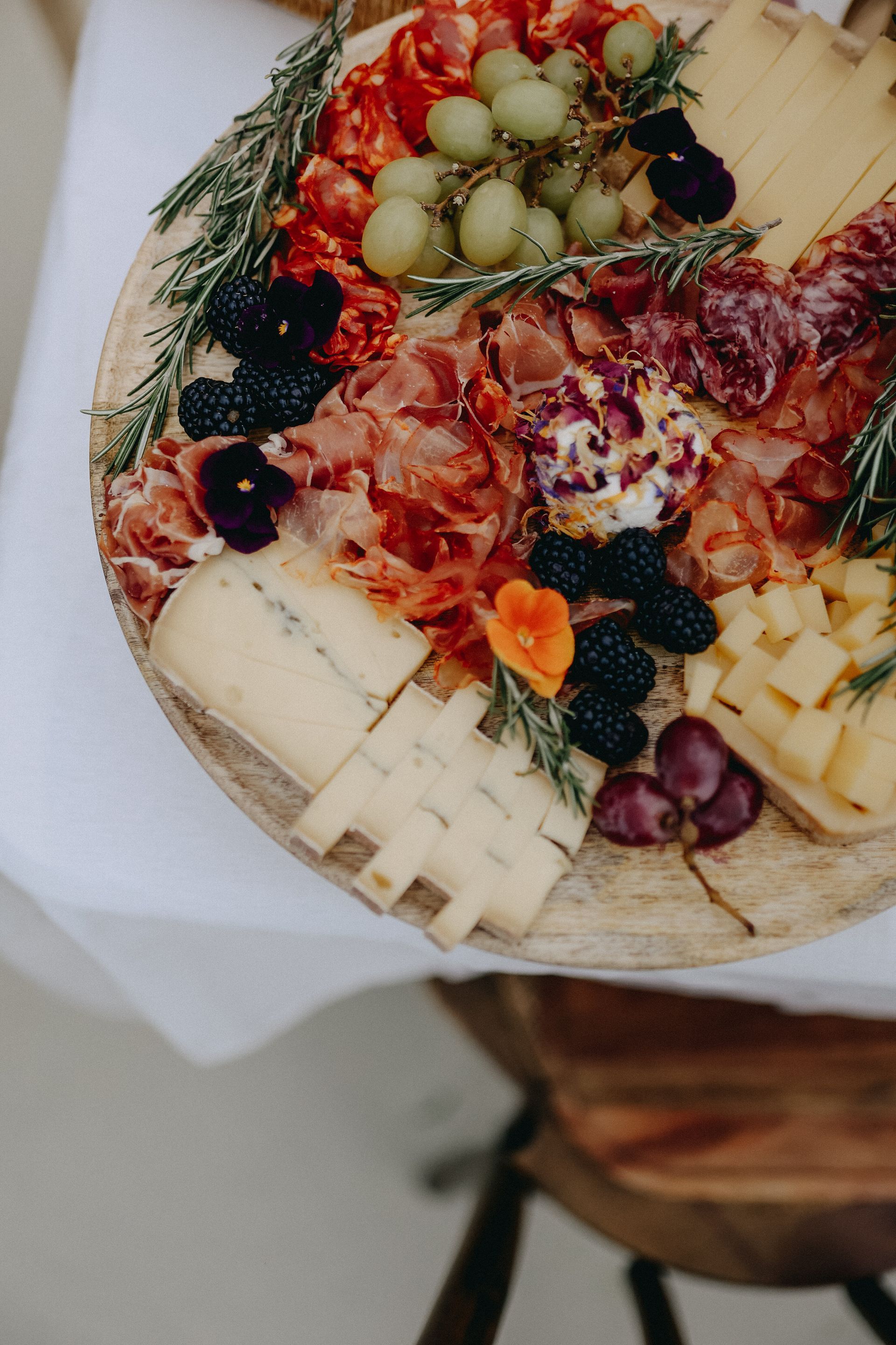 Un plateau en bois garni de fromages variés, de charcuterie, de raisins frais, de mûres et de fleurs comestibles.