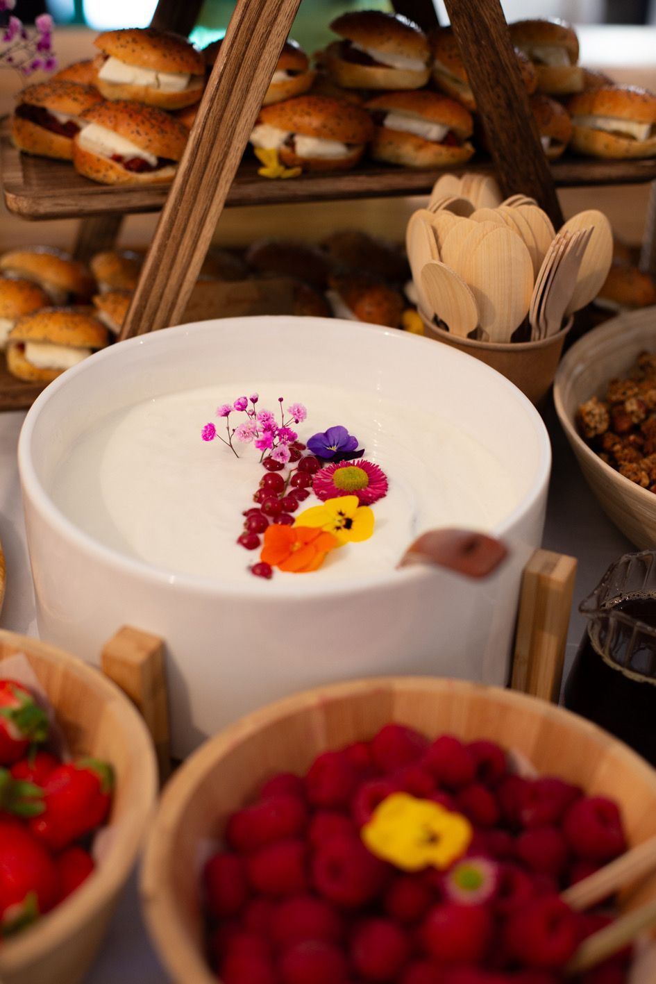 Un buffet de petit-déjeuner avec un bol de yaourt garni de fleurs, des cuillères en bois, des baies et un plateau de petits sandwichs.