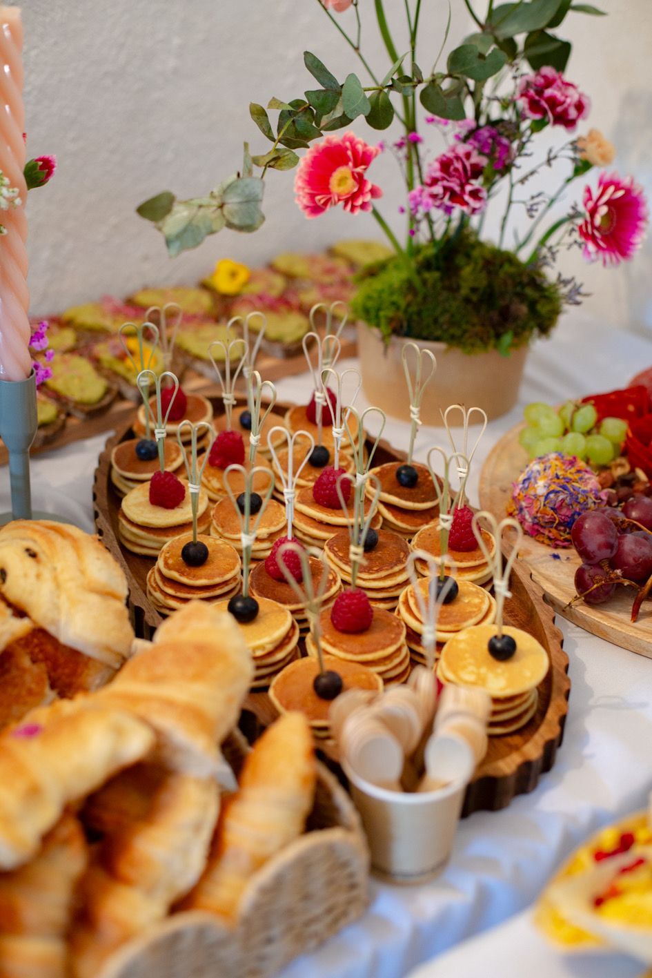 Un buffet festif proposant des mini-brochettes de crêpes garnies de baies, des croissants, des raisins et un centre de table floral éclatant.
