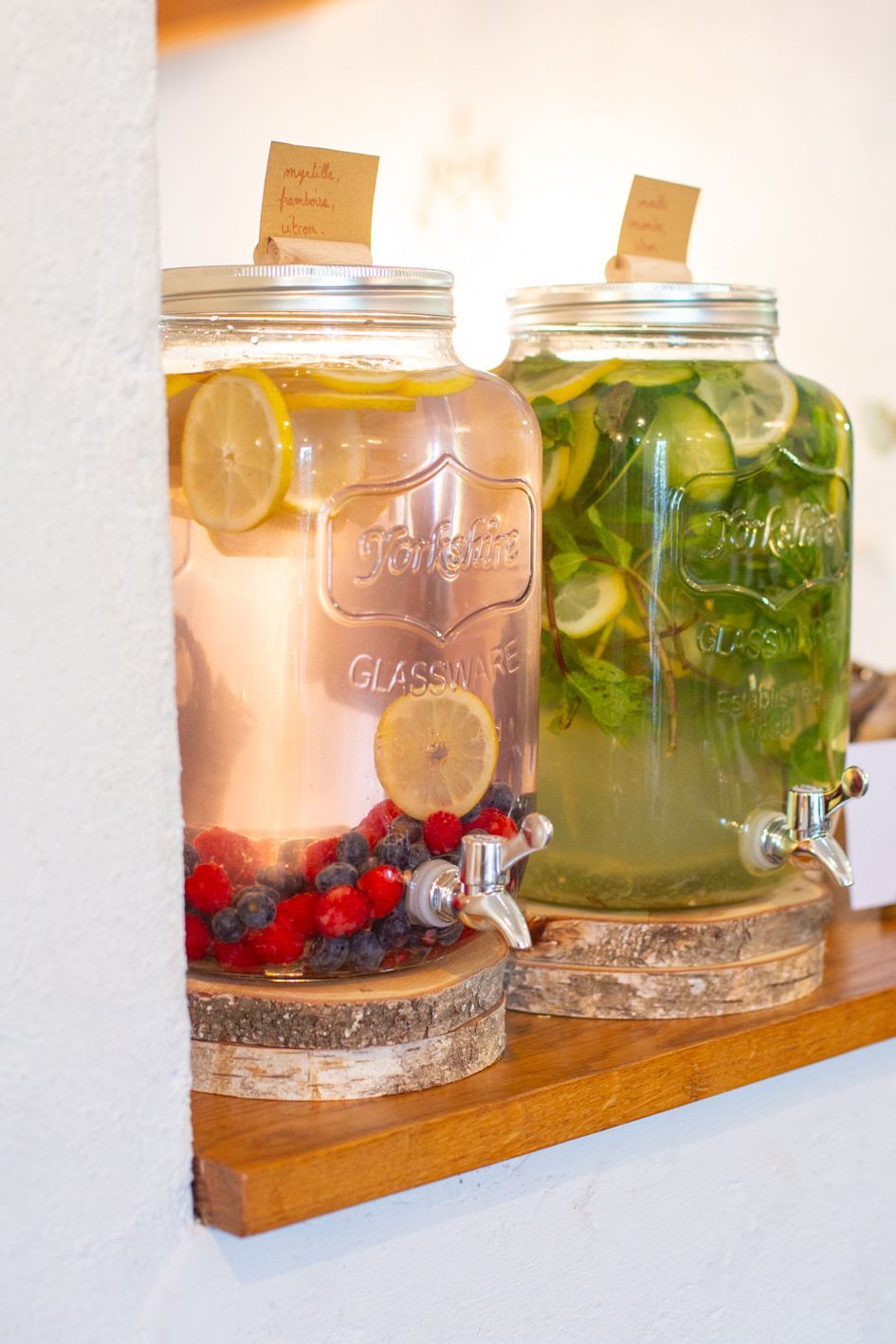 Deux distributeurs de boissons en verre sur des supports en bois, l'un rempli d'eau aromatisée aux fruits, l'autre d'eau au concombre et à la menthe.