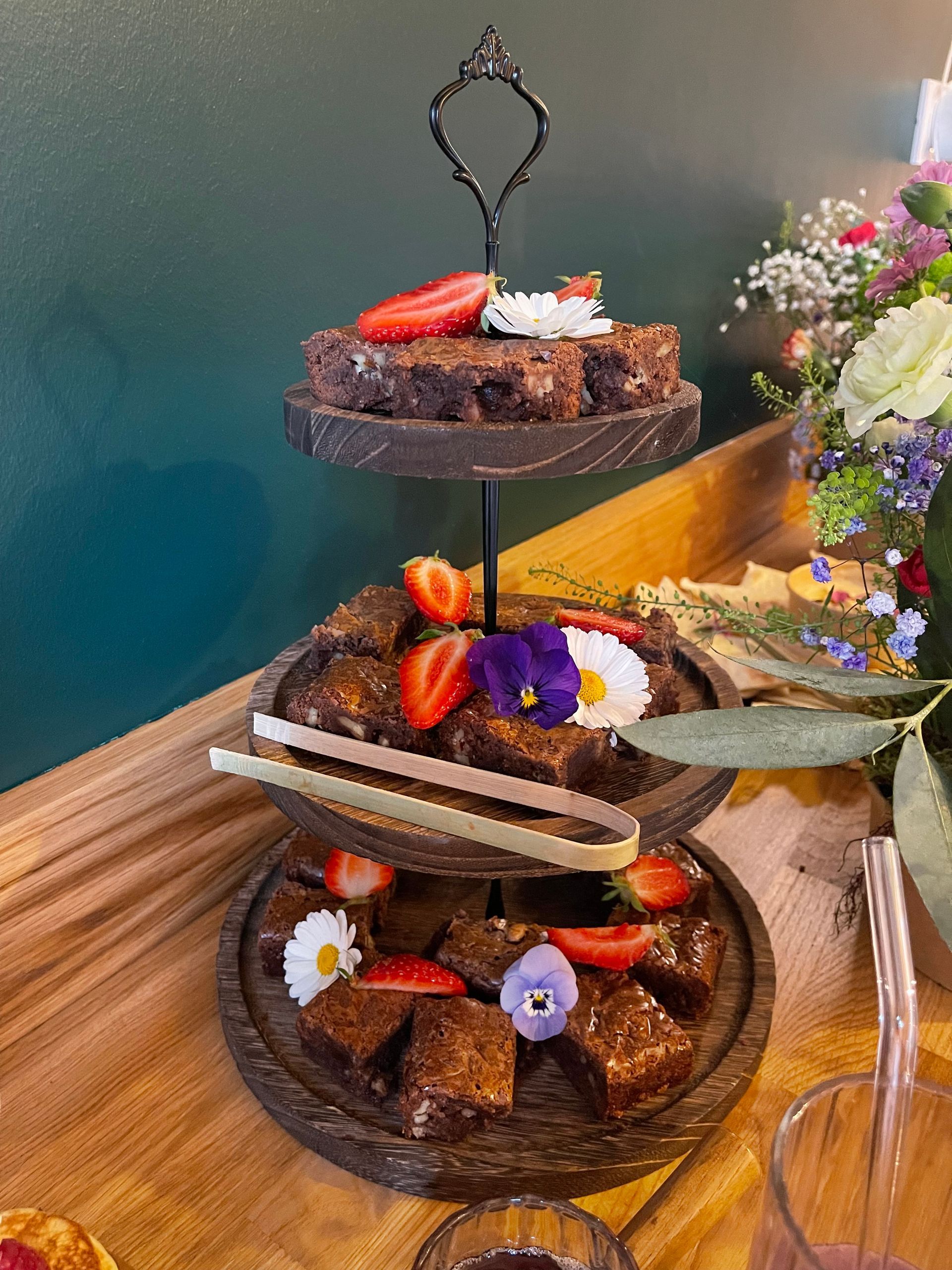 Un plateau de service en bois à trois étages garni de brownies au chocolat, de fraises fraîches et de petites fleurs.