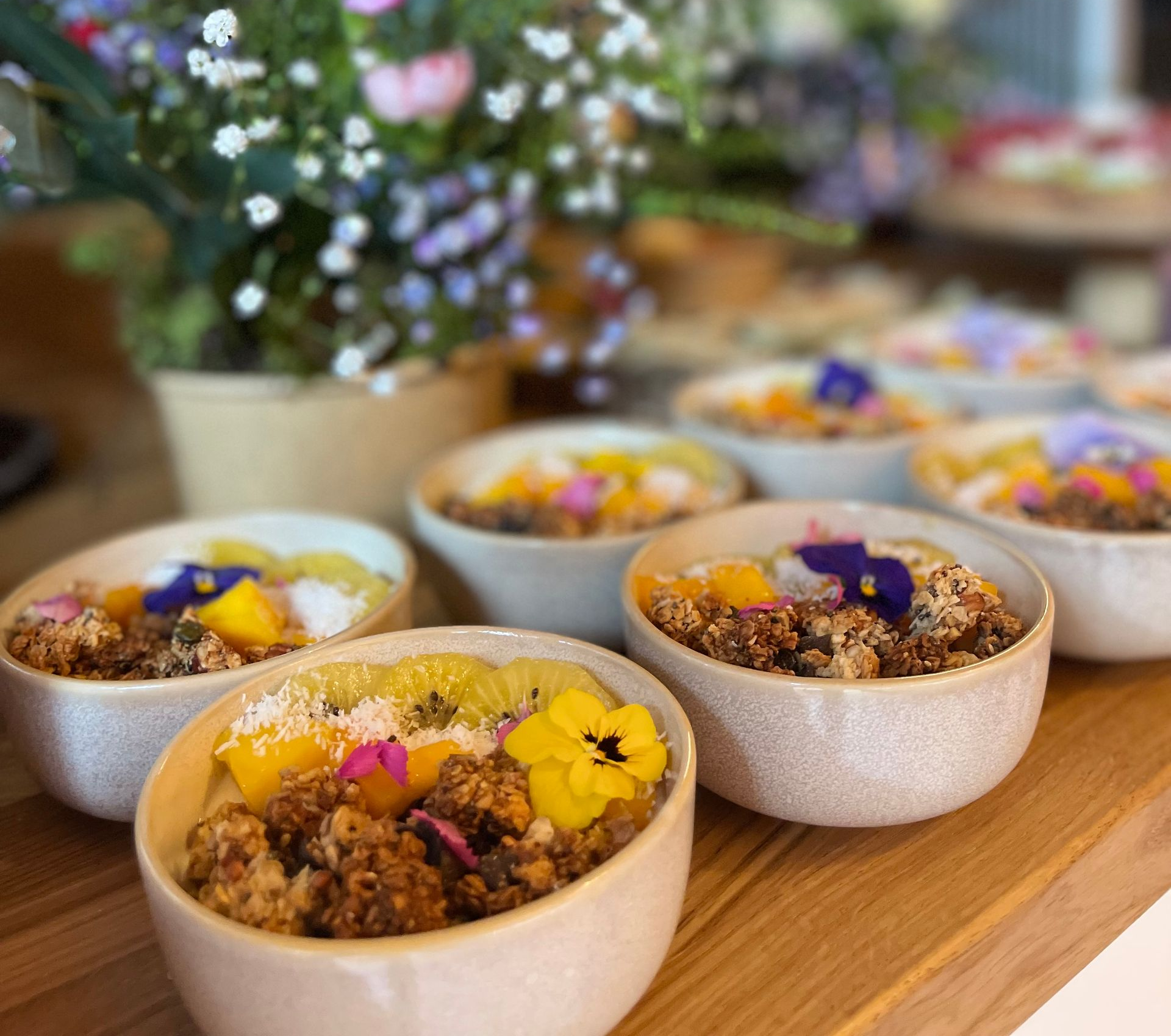 Plusieurs petits bols remplis de granola, de fruits frais et de fleurs comestibles sont disposés sur une surface en bois avec un vase à fleurs.