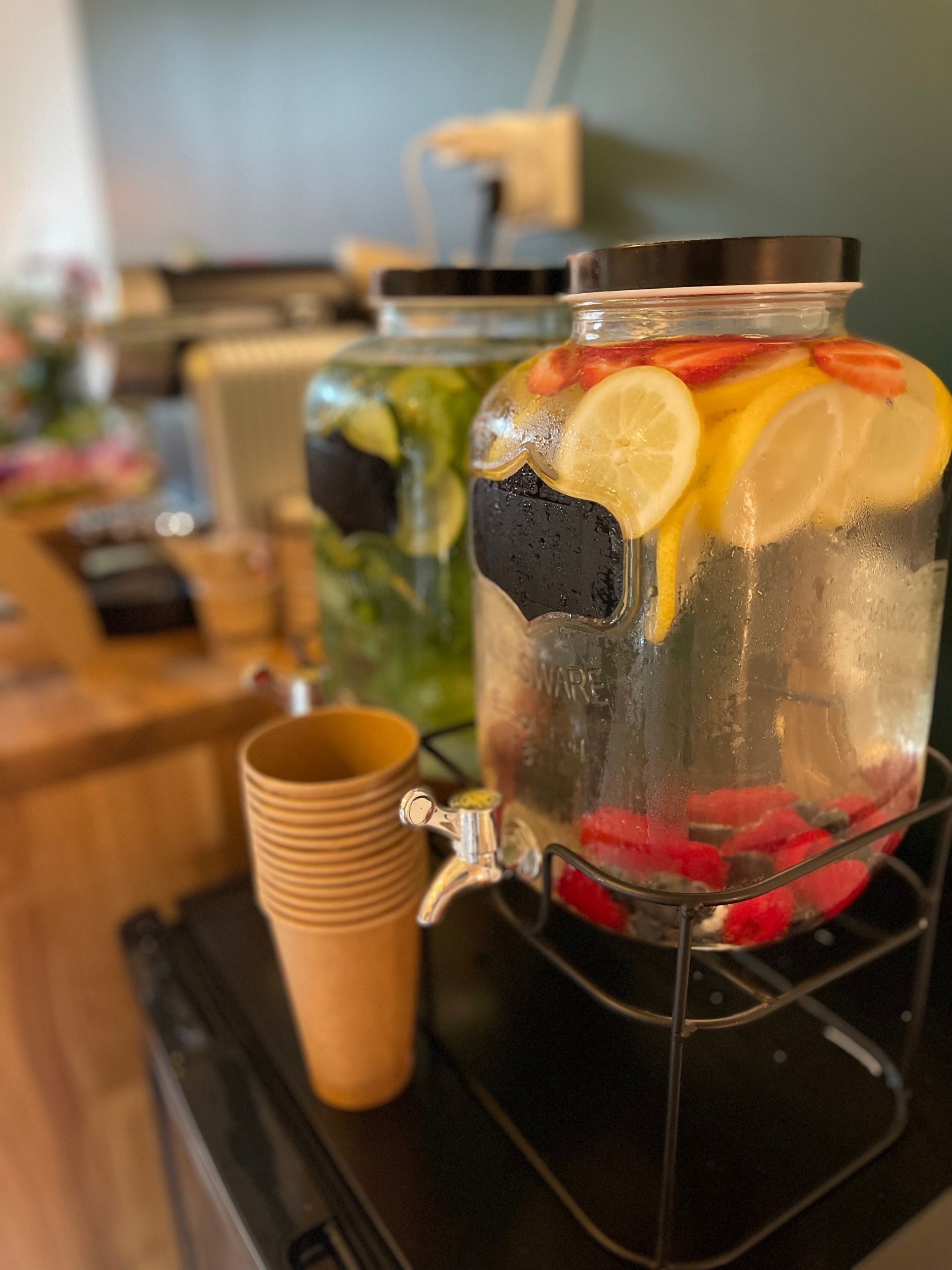 Deux grands distributeurs en verre d'eau aromatisée aux fruits sont posés sur un comptoir à côté d'une pile de gobelets en papier.
