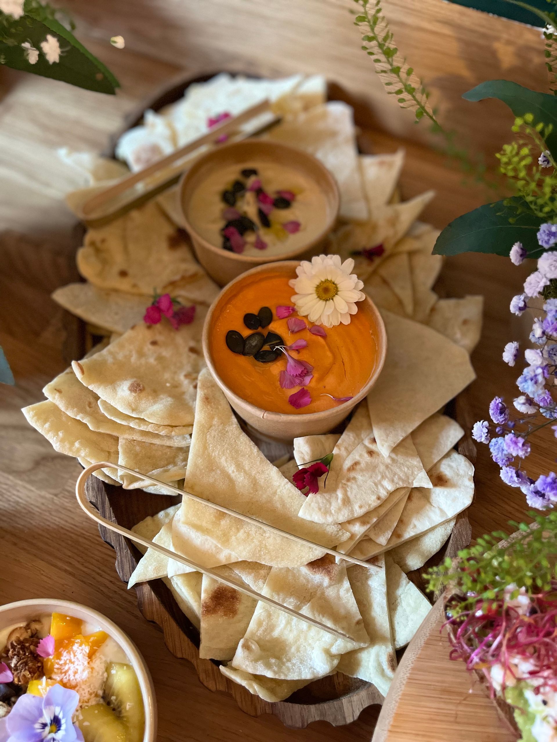 Deux bols de houmous agrémentés de fleurs comestibles et d'olives, entourés de triangles de pain pita sur une surface en bois.