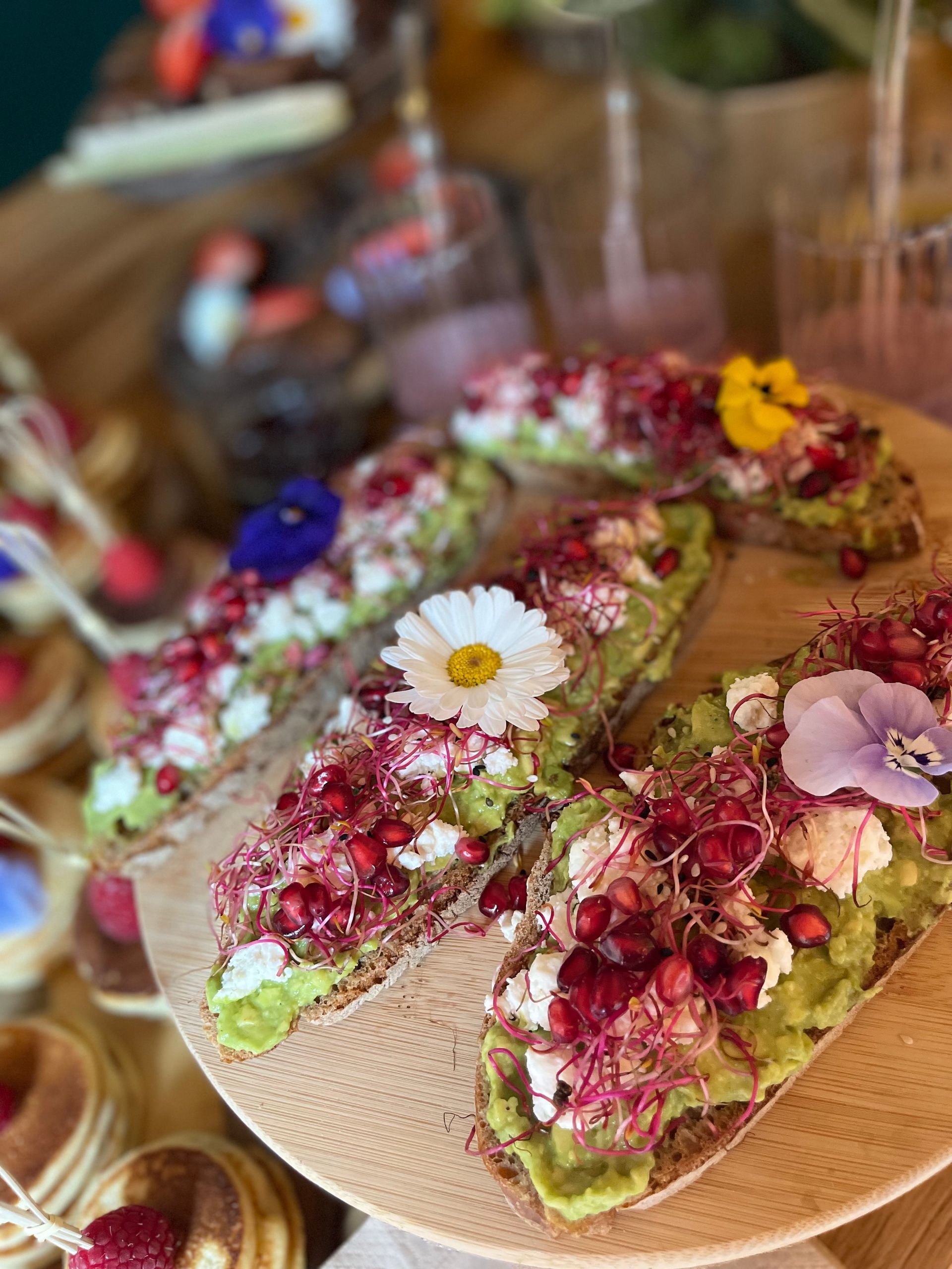 Gros plan sur une tartine d'avocat garnie de graines de grenade, de germes et de fleurs comestibles, présentée sur une planche de service ronde en bois.