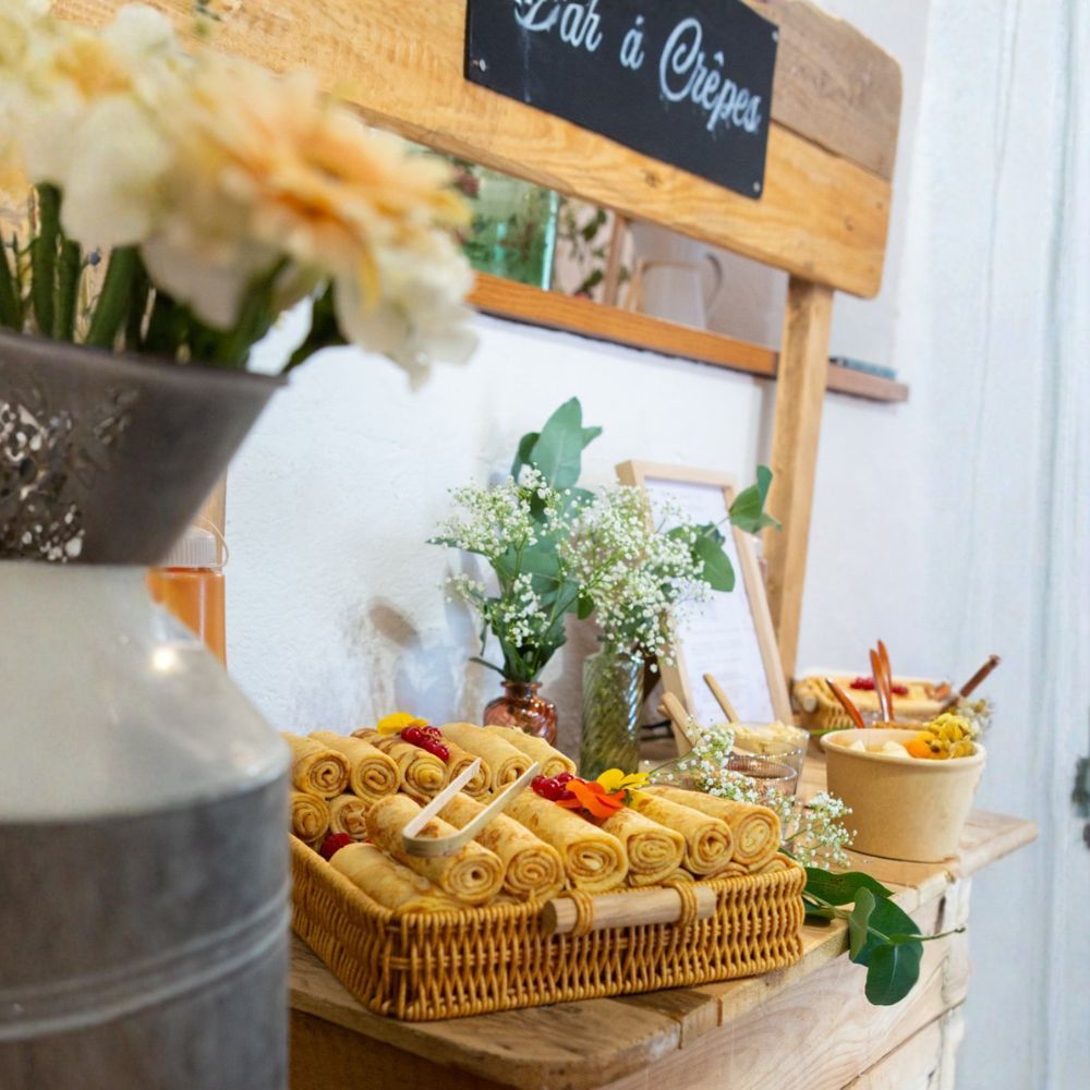 Un présentoir à crêpes comprend un panier en osier rempli de crêpes roulées, un vase à fleurs et un bol de garnitures sur une table en bois.