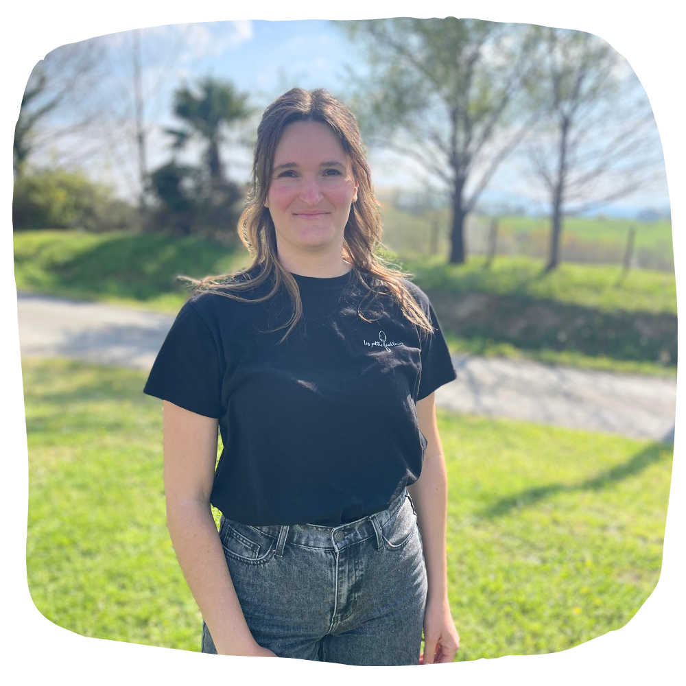 Une personne aux cheveux bruns mi-longs sourit, vêtue d'un t-shirt noir et d'un jean, dans un cadre extérieur ensoleillé et herbeux.