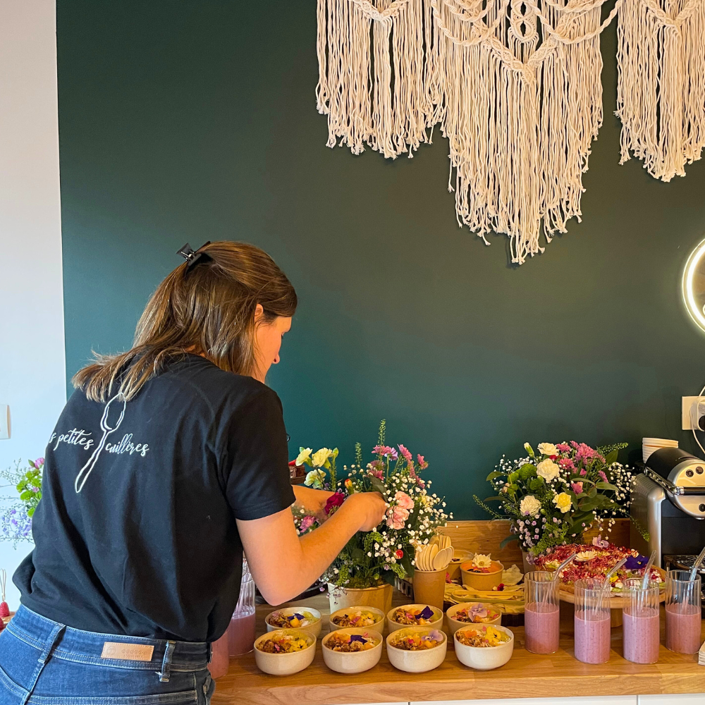 Une personne vêtue d'un t-shirt noir dispose des fleurs sur un comptoir, près de petits bols de snacks et de verres de boissons roses.