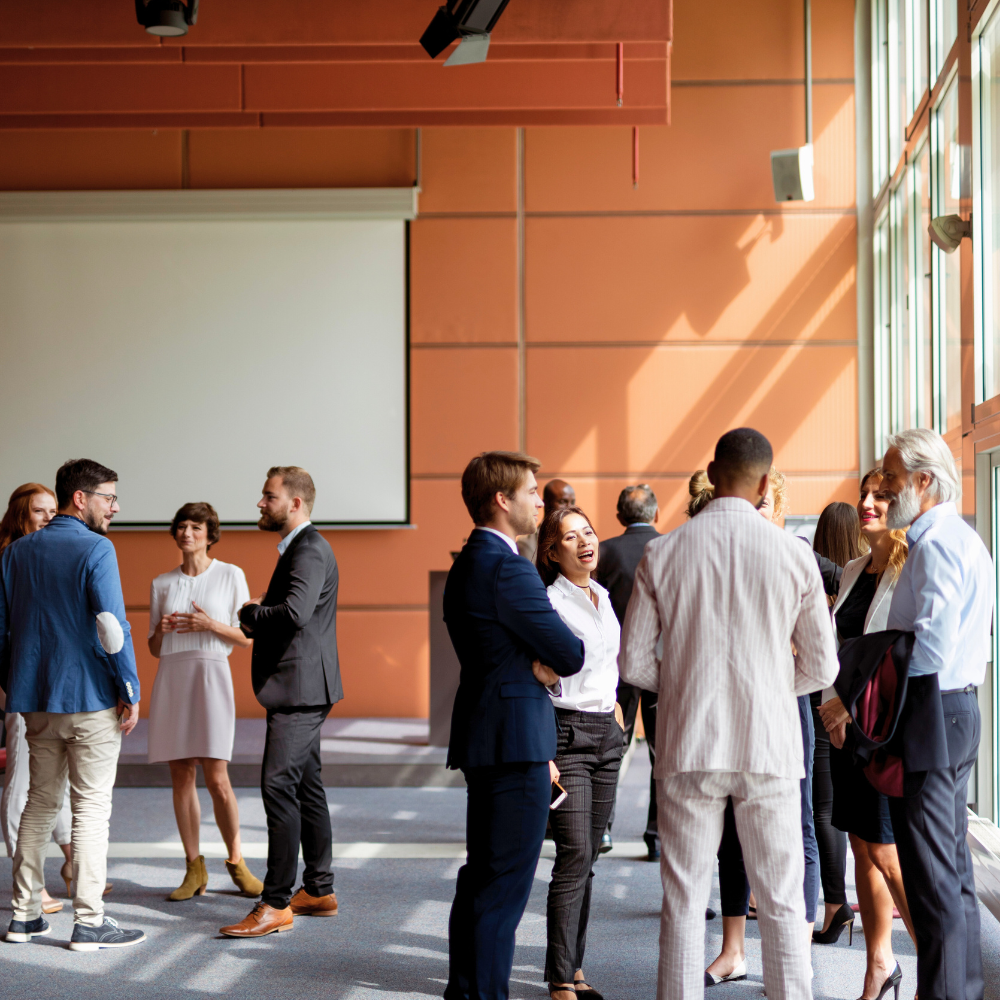 Un groupe de professionnels échange dans une salle moderne et ensoleillée aux murs orange et dotée d'un grand écran de projection vierge.
