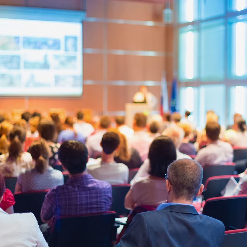 Vue floue depuis l'arrière d'un public regardant un orateur présentant sur un grand écran dans une salle de conférence lumineuse.