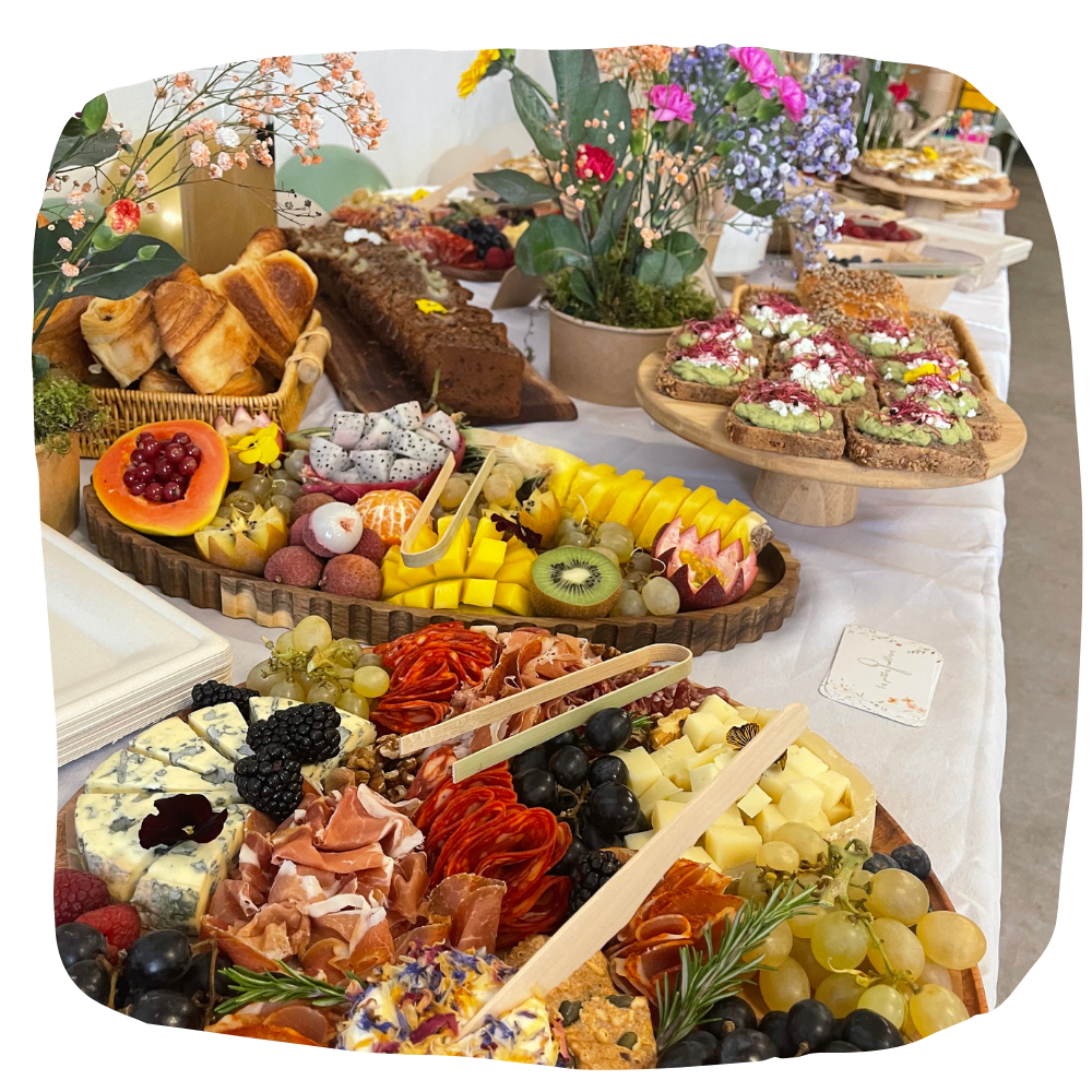 Un assortiment d'amuse-gueules, de fruits, de fromages et de pâtisseries disposés sur des plateaux en bois, sur une nappe blanche.