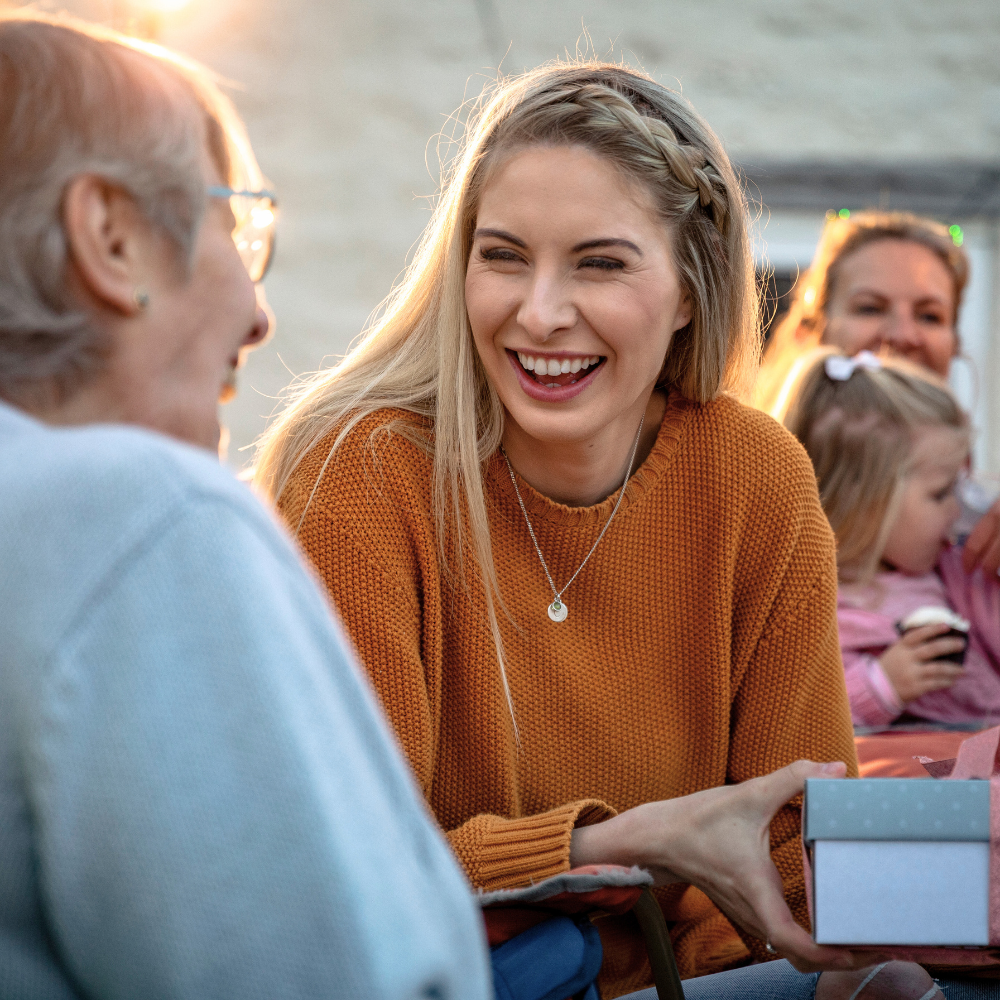 Une personne souriante, vêtue d'un pull orange, tend une petite boîte cadeau à une personne assise en face d'elle, à l'extérieur.