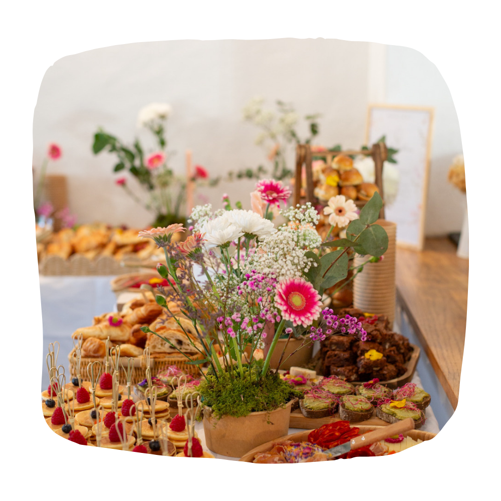 Le buffet traiteur comprend un centre de table floral, des plateaux de pâtisseries, des brownies, des amuse-gueules et des petits canapés.