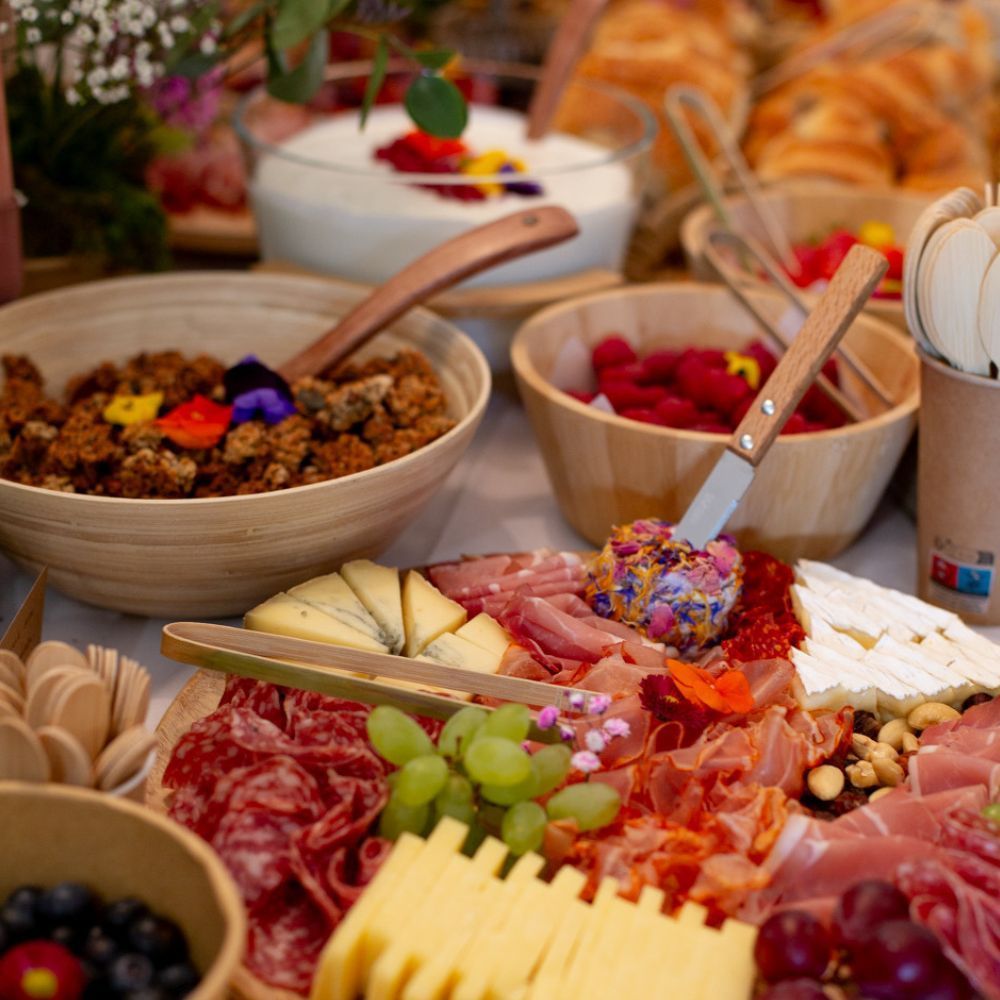 Un petit-déjeuner composé de yaourt, de granola, de fruits frais, de fromages et de charcuterie, disposé sur une table.