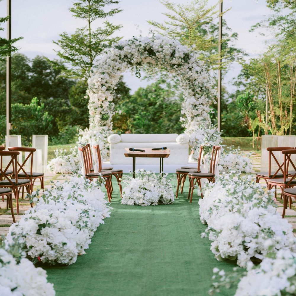 Un décor luxuriant pour une cérémonie de mariage en plein air, avec une arche recouverte de fleurs, une allée tapissée de moquette verte et des rangées de chaises en bois.