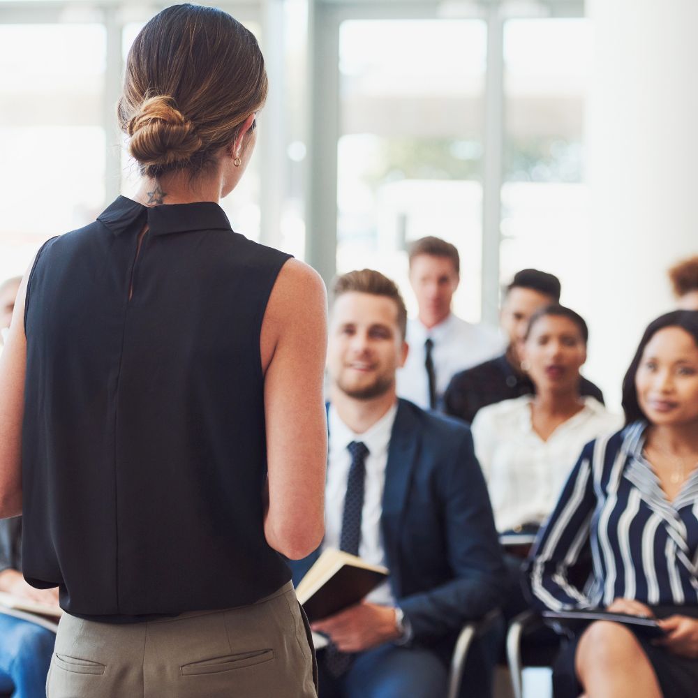 Un présentateur se tient devant un petit groupe de collègues attentifs, assis dans un bureau moderne et lumineux.