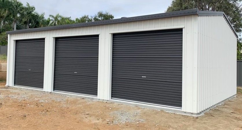 A white garage with three black garage doors is sitting on top of a dirt field.