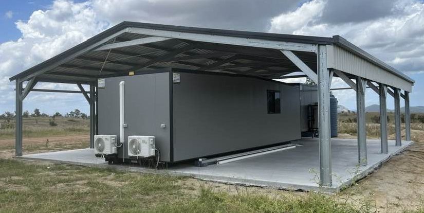 A gray trailer is parked under a metal structure in a field.