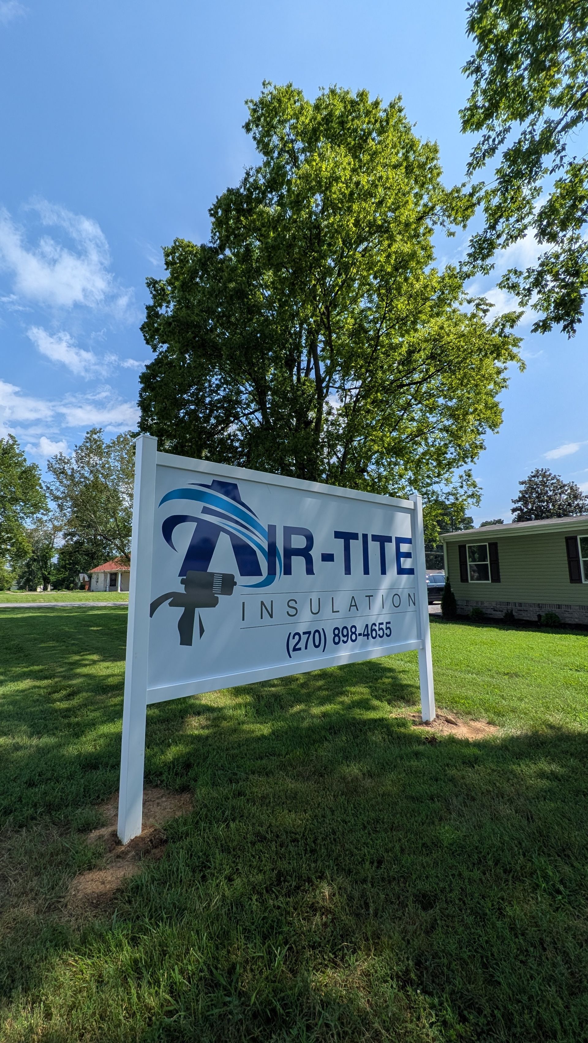 Sign for Air-TITE Insulation. White with logo, blue text, and phone number in front of a green tree.