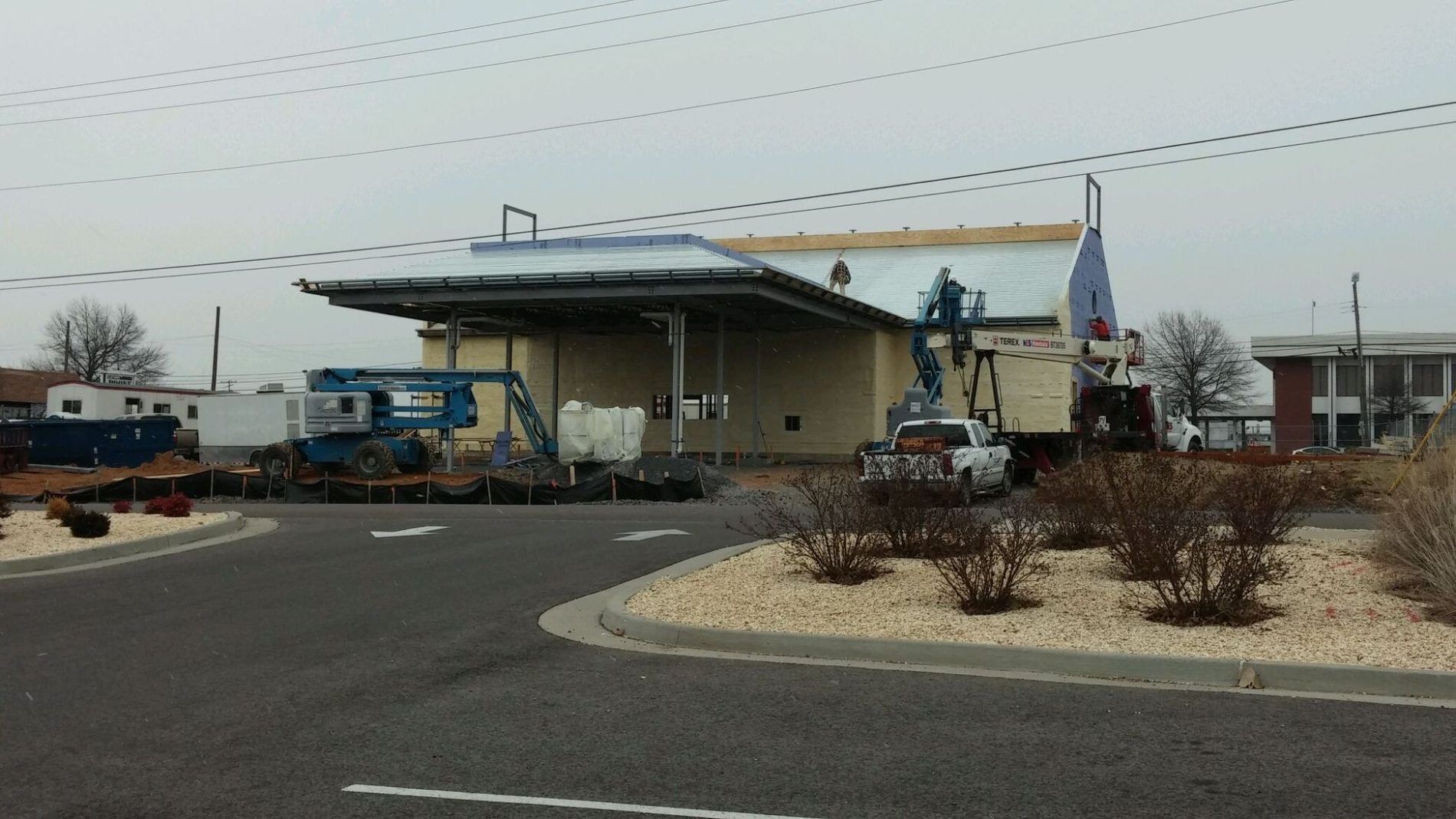 A facility under construction with construction vehicles, a curved driveway, and shrubbery. Overcast day.