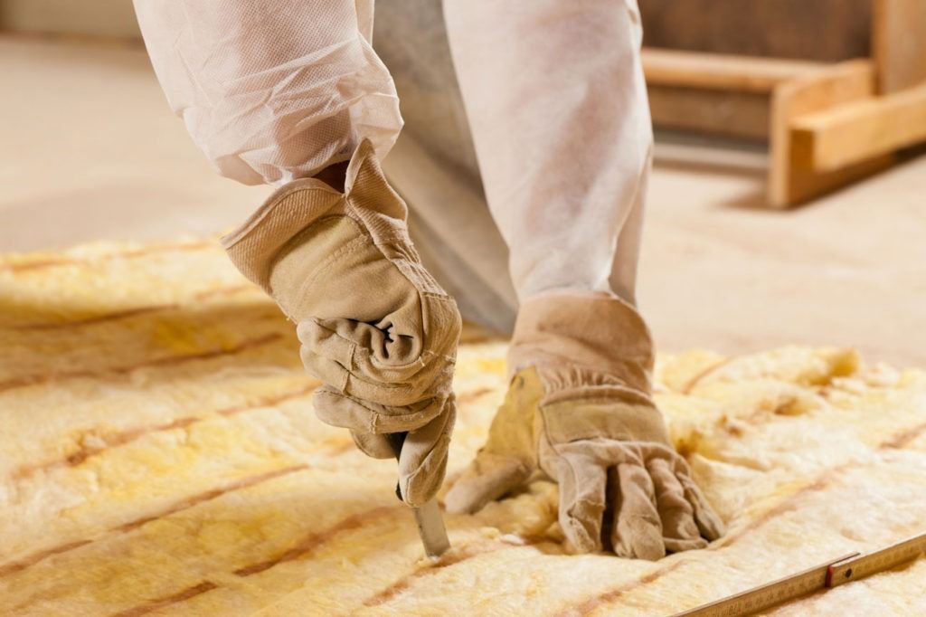 Person cutting insulation with a utility knife, wearing gloves and protective suit indoors.