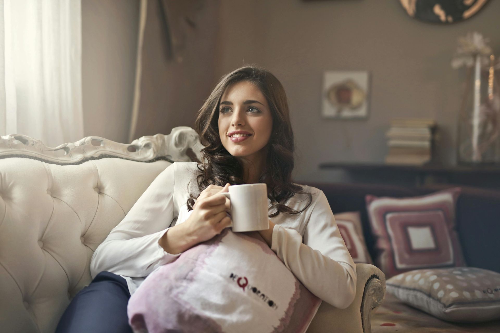 Woman on sofa holding mug, looking out window, smiling. Neutral background, sunlight.