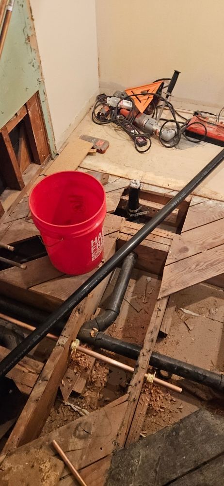 A red bucket is sitting on top of a wooden floor next to pipes.