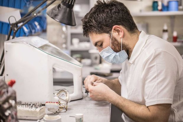 Man in a lab coat, mask, working on a dental mold with tools at a dental lab workspace.