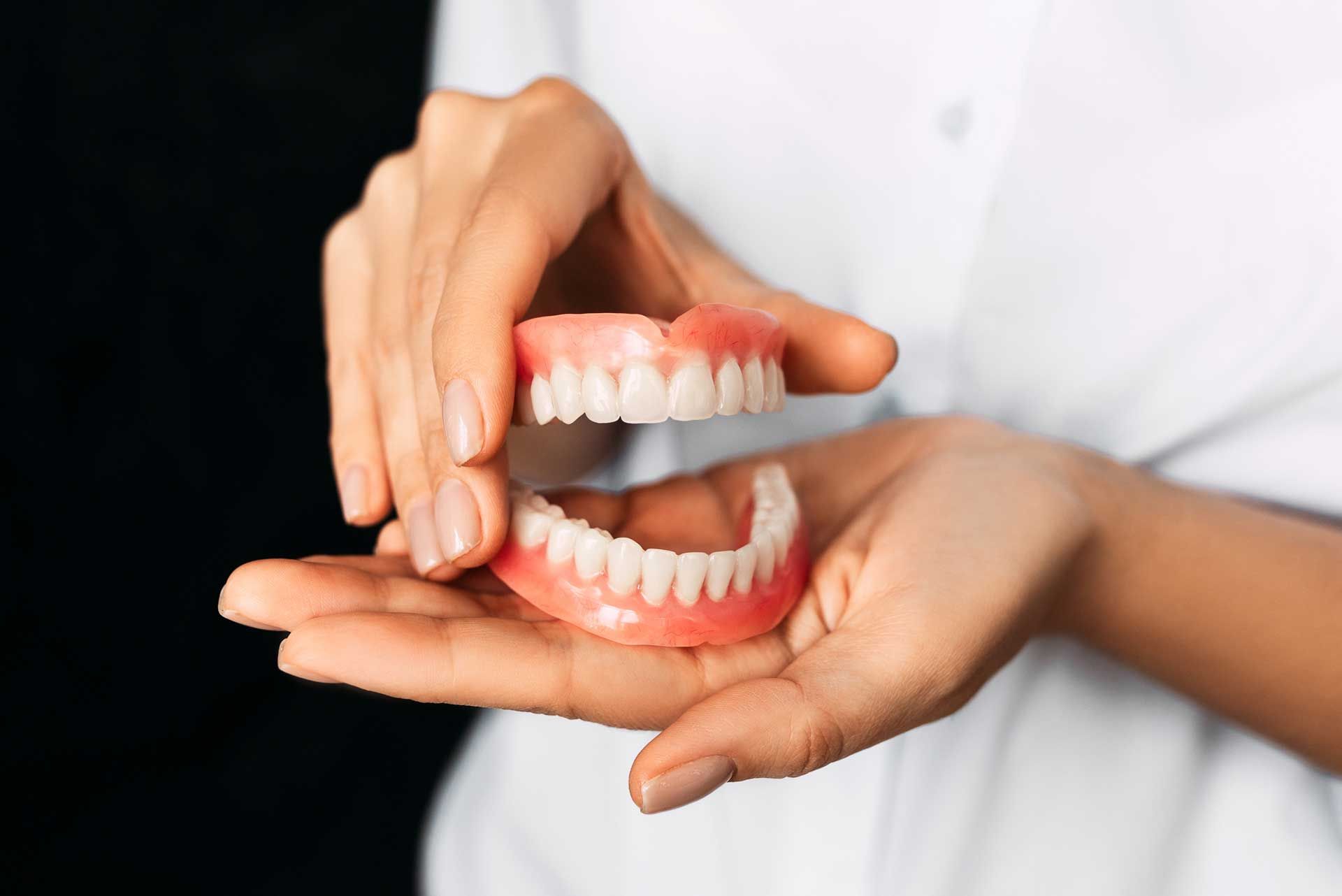 Hands holding upper and lower dentures, pink gums and white teeth, against a dark background.