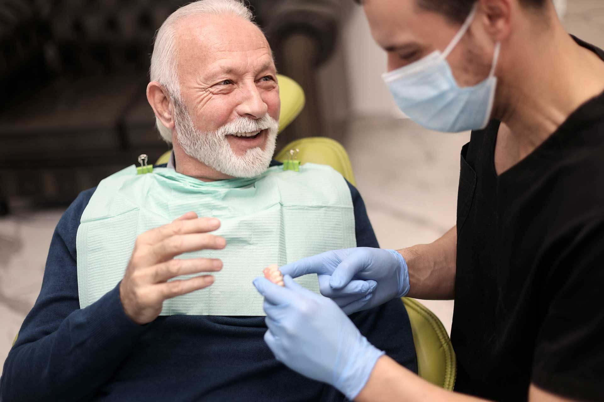 Man in dental chair smiles at dentist holding a tooth sample.