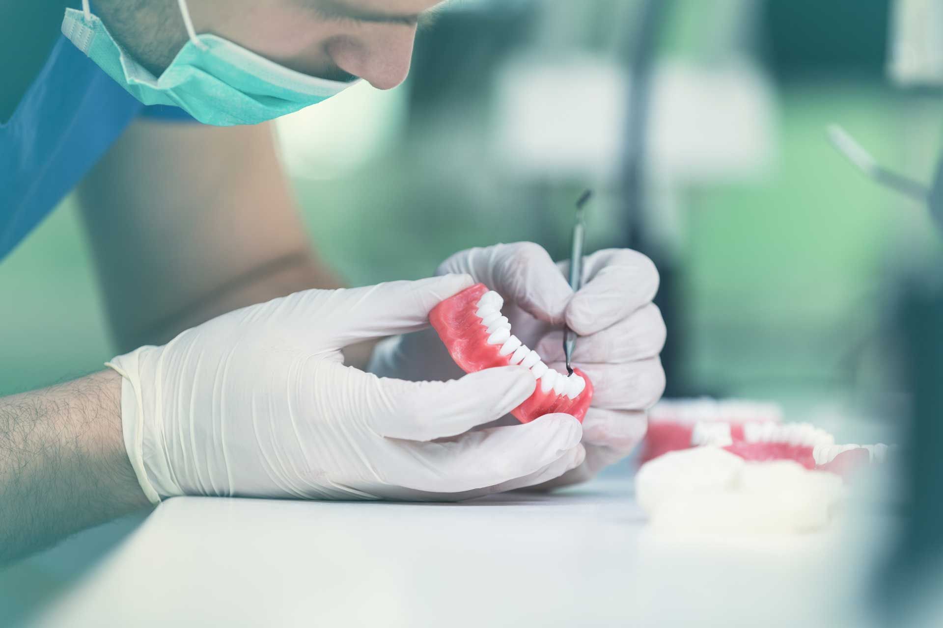 Dentist working on a set of dentures. He wears gloves, a mask, and uses a tool in a lab setting.