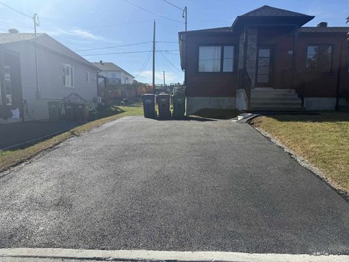 Une allée asphaltée fraîchement pavée mène à une maison brune avec des marches, flanquée d'une maison voisine et de poubelles.