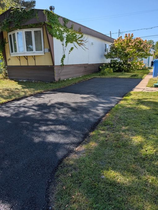 Une allée en asphalte sombre fraîchement pavée mène à une maison mobile recouverte de vigne sous un ciel radieux et ensoleillé.