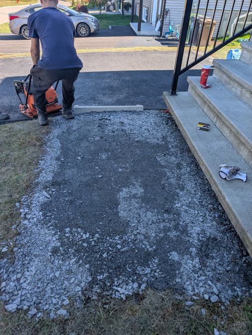 Une personne utilise une plaque vibrante sur une base de gravier à côté de marches en béton lors d'un chantier de construction extérieur.