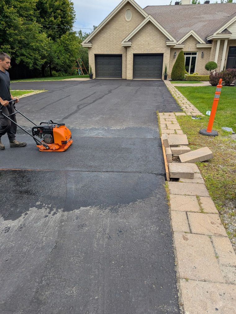 Un ouvrier utilise une plaque vibrante orange pour aplanir l'asphalte frais d'une allée devant une maison.