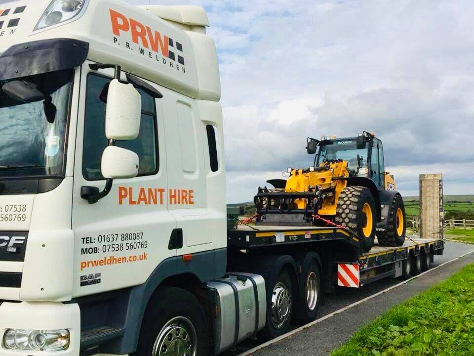 a plant hire truck is carrying a tractor down a road .