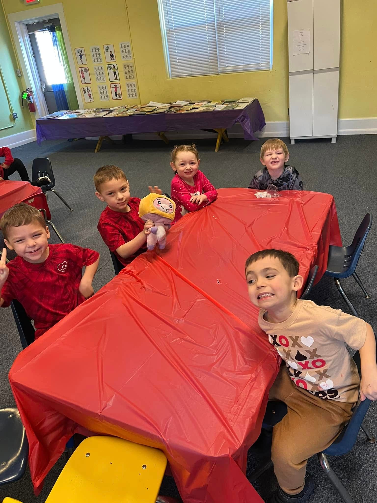 A group of children are sitting at a table with a red table cloth.