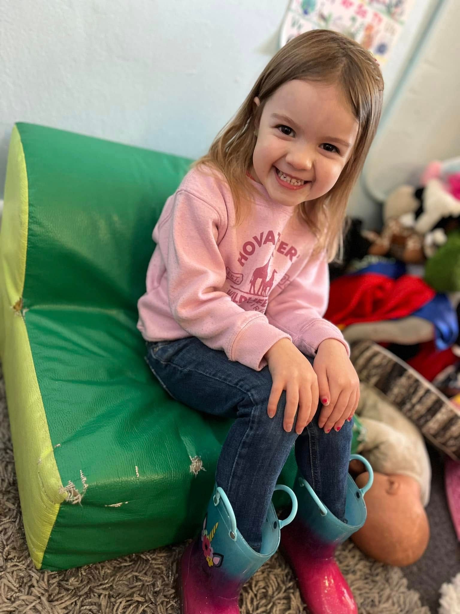 A little girl is sitting on a green couch wearing pink boots.