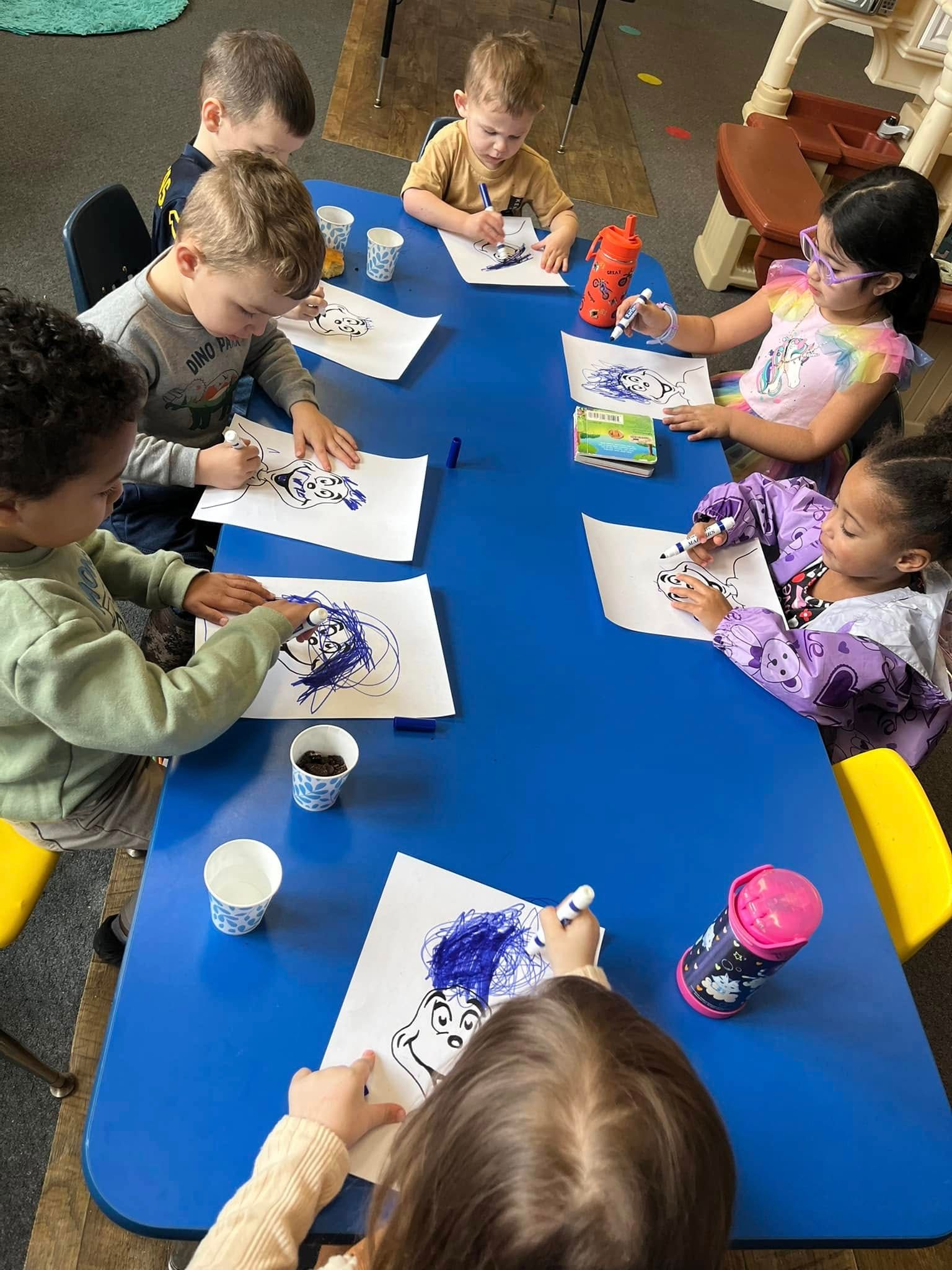 A group of children are sitting at a blue table drawing.