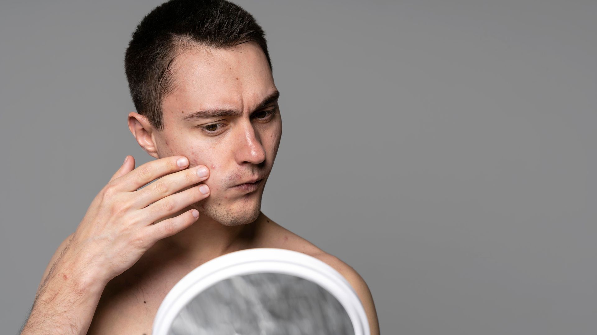 A shirtless man is looking at his face in a mirror.