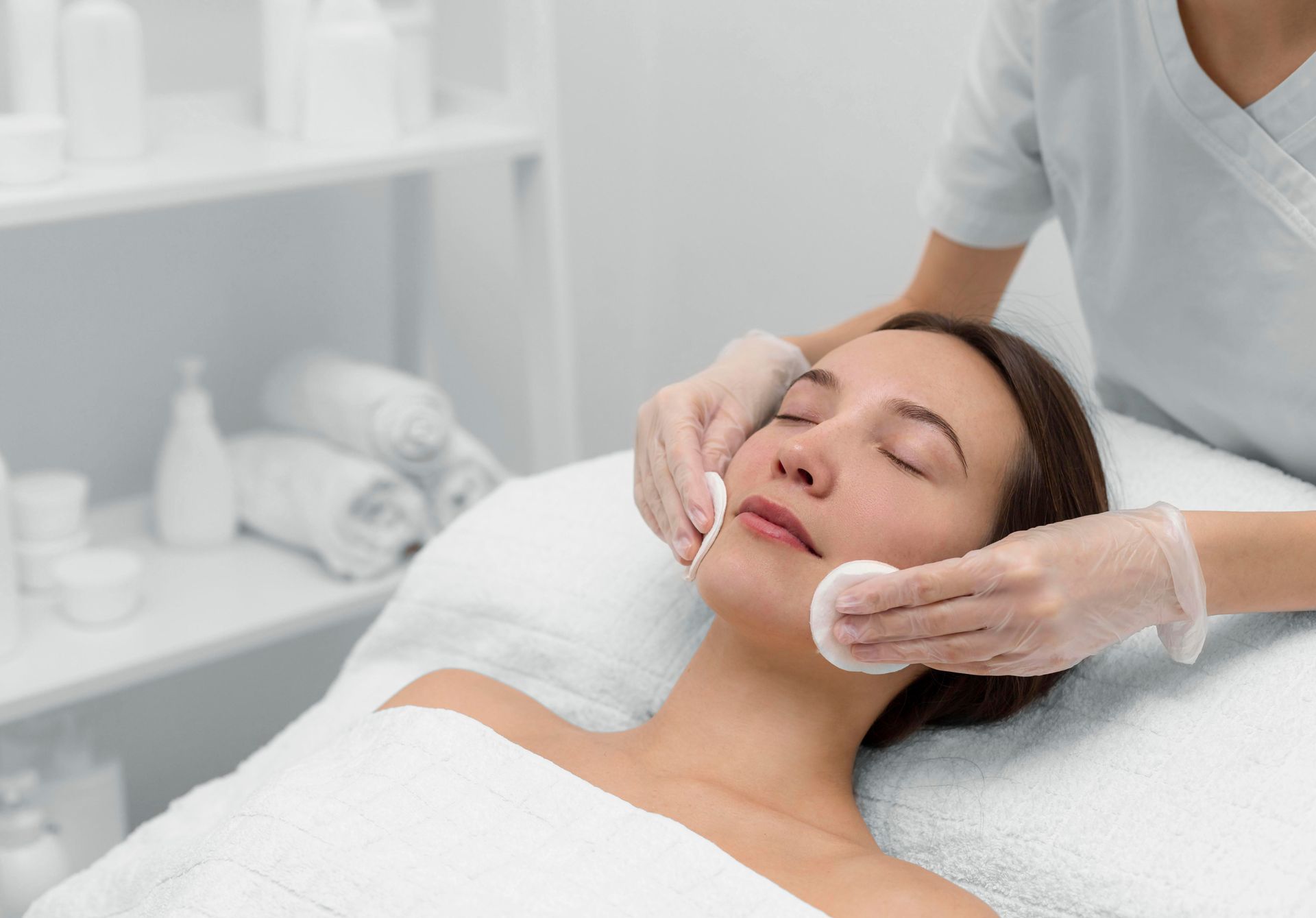 Woman getting facial at a spa. Technician uses cotton pads on her face. White linens, neutral background.